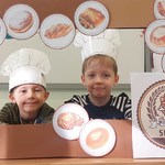 Dwóch chłopców w kucharskich czapkach pozują do zdjęcia w ramcę do której przyklejone są chleby.jpg