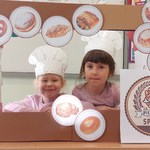 Dwie dziewczynki w kucharskich czapkach pozują do zdjęcia w ramcę do której przyklejone są chleby.jpg